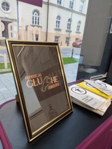 Zbliżenie na stół przy oknie, na którym stoi w czarnej ramce grafika ze złotym napisem „Fundacja Głuche Miasto”, z charakterystyczną literką „C” w języku migowym. Obok leży stos kolorowych książeczek, głównie białych i żółtych, z symbolem dostępności i hasłem „Jak korzystać z prawa do dostępności”. W tle przez szybę widać fragment chodnika, ulicy i zabytkowego budynku z białą elewacją oraz zaparkowany czerwony samochód