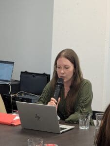 Kobieta z długimi brązowymi włosami i okularami mówi do mikrofonu, siedząc przy ciemnym stole z otwartym laptopem HP. Ubrana jest w ciemnozieloną sukienkę z marszczeniami na rękawach. Przed nią leżą dokumenty w czerwonej teczce oraz szklanka wody. W tle widoczny jest drugi laptop, pętla indukcyjna oraz biało-szara ściana. W prawym dolnym rogu kadru widoczna część głowy innej osoby