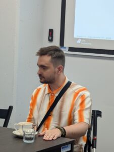 Mężczyzna z krótkimi, ciemnymi włosami i zarostem siedzi przy stole, opierając dłoń na jego blacie. Ubrany jest w koszulę w pomarańczowo-białe pasy i ma zawieszoną na ramieniu czarną torbę. Na nadgarstku nosi bransoletki, a obok jego krzesła stoi kula ortopedyczna. Na stole przed nim stoją filiżanka ze spodkiem oraz szklanka wody. W tle widać fragment ekranu z informacją o pogodzie: 22°C, pochmurnie