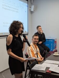 Młoda kobieta z burzą czarnych loków, z mikrofonem w ręku przemawia stojąc przy ciemnym stole. Ubrana jest w czarną sukienkę z wycięciem na ramieniu i trzyma kartkę z tekstem. Obok niej siedzi mężczyzna w koszuli w pomarańczowo-białe pasy, uśmiechający się w stronę aparatu. W tle na pudełku z logo Lublina i napisem „Miasto Młodych” siedzi inny mężczyzna – tłumacz języka migowego - ubrany na czarno, pokazujący znak w języku migowym. Za nimi znajduje się ekran z wyświetlonymi napisami z dokumentu edytowanego na żywo w programie WORD