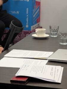 Zdjęcie przedstawia ciemny stół z rozłożonymi dokumentami, w tym widocznymi certyfikatami z logo PFRON, RPO i Fundacji Głuche Miasto. Na stole znajdują się także telefon w czerwonym etui, dwie szklanki z wodą, biała filiżanka z resztką kawy na spodku oraz zamknięty laptop. W tle kolorowe pudełka z napisem „Miasto Młodych” oraz hashtagiem Lublin is Youth. Po lewej stronie widoczna część kul ortopedycznych i siedząca osoba w ciemnym ubraniu