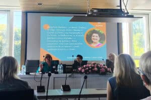 Trzy kobiety prowadzące spotkanie, siedzą za stołem konferencyjnym. Wśród nich prof. Barbaro, której zdjęcie i wyświetla się na monitorze wraz z krótką notatką o badaczce