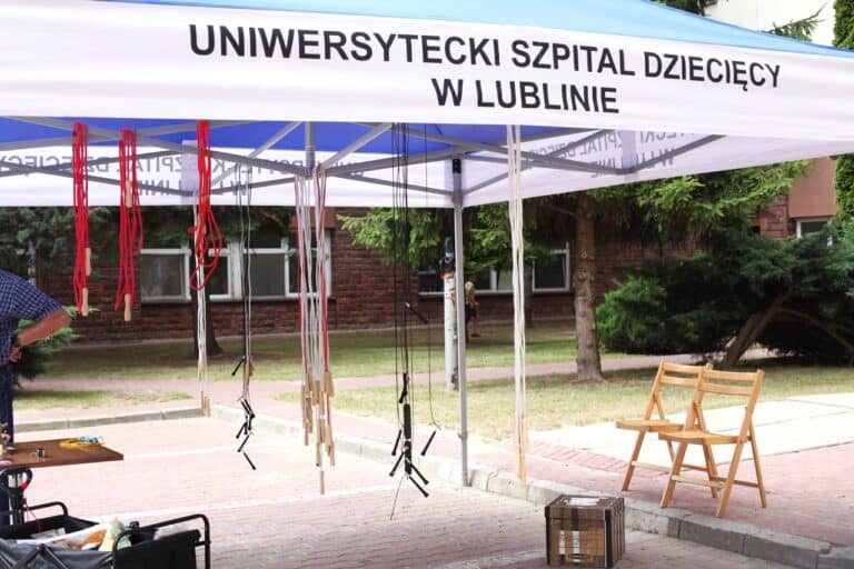 Widok na fragment namiotu z napisem Uniwersytecki Szpital Dziecięcy w Lublinie. W centralnej części kadru, na konstrukcji namiotu zawieszono kolorowe skakanki