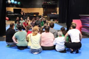 Wnętrze sali z lustrem wypełniającym niemal całą ścianę. W lustrze odbicie grupy uczestników warsztatów, którzy siedzą na niebieskiej macie i patrzą na siebie w lustrze