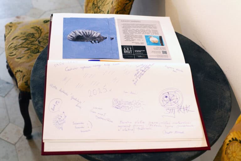 Widok na karty księgi pamiątkowej z wklejonymi ulotkami informującymi o wystawie Zwierzęta znane i nieznane Adama Sarana. Na karcie wpisy gości i gratulacje