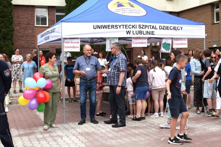 Widok na Punkt Liczenia Uczestników - namiot z napisem Uniwersytecki Szpital Dziecięcy w Lublinie, z logo placówki. Wokół namiotu tłoczą się uczestnicy wydarzenia. Charakterystyczna jest kobieta z personelu medycznego uchwycona w lewej części kadru, trzymająca w ręku różnokolorowe balony