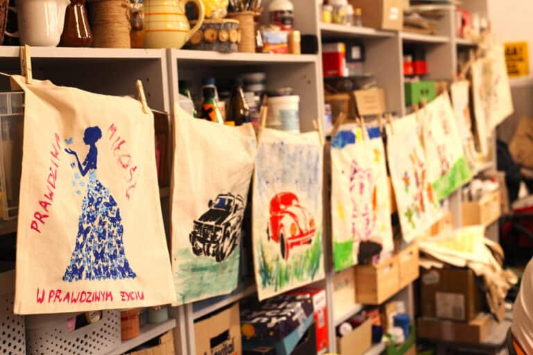 Zbliżenie na ekspozycję pomalowanych przez dzieci toreb. Torby przyczepiono do sznurka drewnianymi spinaczami, wzdłuż regału z akcesoriami plastycznymi. Przypominają rozwieszone pranie. Ostrość w kadrze skoncentrowana jest na pierwszej torbie z wizerunkiem kobiety w niebieskiej sukni z motyli i czerwonym napisem: „Prawdziwa miłość w prawdziwym życiu”