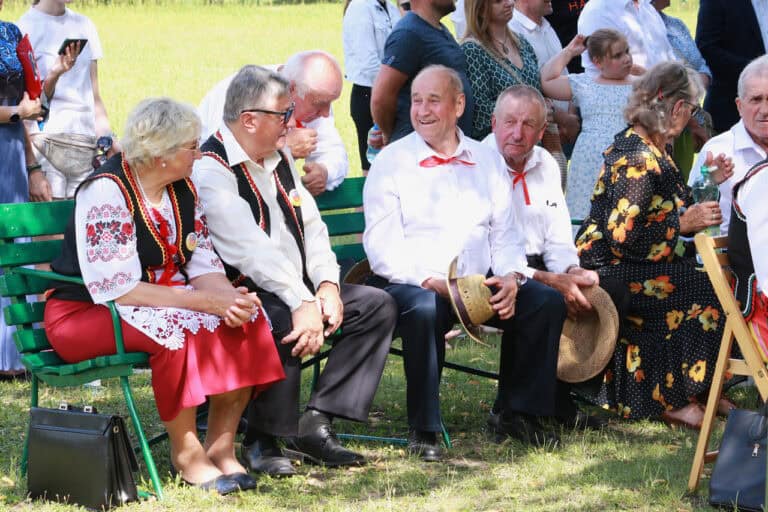 Publiczność. Zbliżenie na grupę seniorów: czterech mężczyzn i jedną kobietę. Pogrążeni w rozmowie patrzą na śmiejącego się mężczyznę. Wszyscy w odświętnych kolorowych strojach: białych koszulach i ciemnych spodniach. Kobieta w białej haftowanej bluzce, czerwonej spódnicy i obszytej haftem czarnej kamizelce
