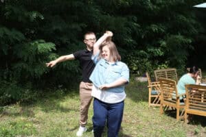 Na tle zieleni para taneczna podczas wykonywania obrotu. Oboje uśmiechają się. Z prawej strony kadru fotele ogrodowe, w jednym z nich siedzi jedna osoba