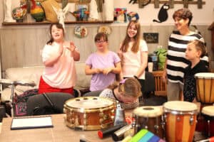 Na pierwszym planie instrumenty muzyczne (głównie bębny). Drugi plan to dzieci i opiekunka. Dzieci uśmiechają się i klaszczą. W tle półka z bibelotami rękodzielniczymi