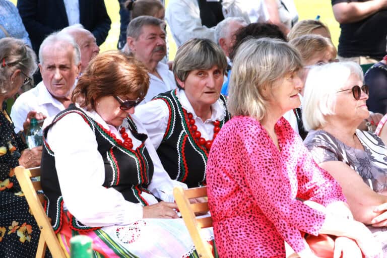 Grupa kilkunastu siedzących osób. Wśród nich kobieta patrząca wprost do obiektywu. Wszyscy w kolorowych odświętnych strojach. Kobieta ma na sobie kolorowy strój i grube czerwone korale