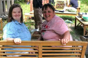 Dwie młode kobiety siedzą na ławce ogrodowej i śmiejąc się patrzą w obiektyw aparatu. Jedna ma na sobie różową koszulkę, a druga niebieską, dżinsową koszulę. W tle krzesła i ławka