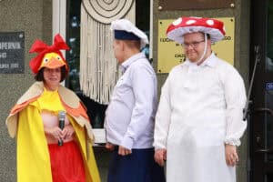 11. Trzy osoby. Od lewej: kobieta w żółto - pomarańczowym stroju kaczki z czerwoną kokardą na głowie, trzyma w rękach mikrofon. Obok niej mężczyzna stoi bokiem do aparatu fotograficznego. Trzecia osoba – mężczyzna, ma na sobie biały fartuch i nakrycie głowy w kształcie kapelusza muchomora – czerwonego w białe duże grochy