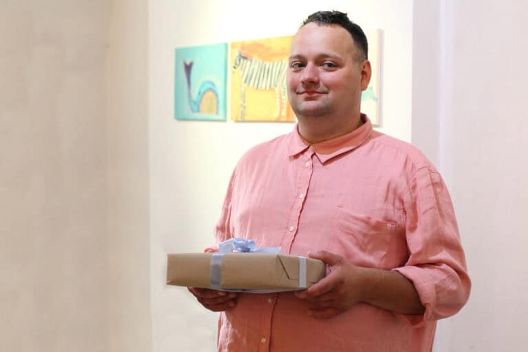 Adam Saran pozujący do fotografii. Artysta jest uśmiechniety, w dłoniach trzyma prezent urodzinowy: pudełko przewiązane błękitną wstążką
