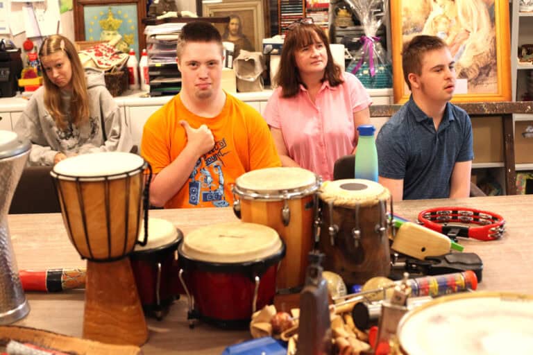 Na pierwszym planie instrumenty muzyczne (bębny, tamburyn, grzechotki) Ostrość na młodzież siedzącą przy stole z instrumentami. Wśród nich opiekunka. Chłopiec, który patrzy wprost do obiektywu unosi w górę kciuk