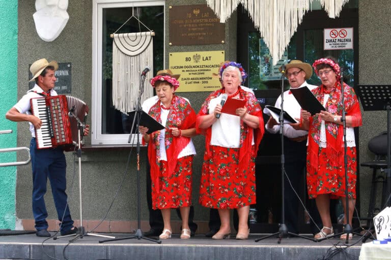 Na scenie grupa siedmiu osób podczas występu seniorów. Od lewej: mężczyzna w kapeluszu, grający na akordeonie, kolejno osoby śpiewające do mikrofonów. Kobiety w kwiecistych strojach, w czerwonych barwach, z wiankami na głowach, mężczyźni w kapeluszach, białych koszulach i ciemnych spodniach