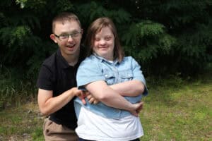 Pozowana fotografia. Na pierwszym planie młoda dziewczyna, za nią młodzieniec w okularach, trzyma ją za ręce. Oboje uśmiechają się patrząc wprost do aparatu. W tle zieleń