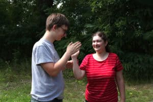 Dwie osoby: nastolatek w szarej koszulce i dziewczyna w czerwonej bluzce w białe poziome paski. Uśmiechają się przybijając sobie „piątkę”. W tle zieleń