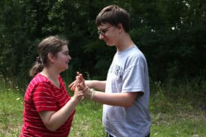 Dwie osoby stoją naprzeciwko siebie na tle zieleni: nastolatek w szarej koszulce i okularach i dziewczyna w czerwonej bluzce w białe poziome paski. Uśmiechają się patrząc na siebie. Trzymają się za ręce