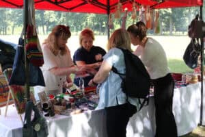 14. Stoisko Warsztatów Terapii Zajęciowej, przy nim cztery kobiety. Dwie z nich prezentują swoje wyroby pozostałym. Podłużna ekspozycja nakryta białym obrusem, a na niej wyroby rękodzielnicze