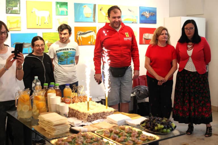 W centralnej części kadru świecące race na torcie urodzinowym otoczonym przez gości. Po lewej stronie kadru kobieta chwyta moment, robiąc zdjęcie aparatem w telefonie