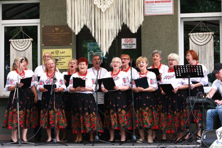 Na scenie grupa seniorów w lokalnych strojach: białe haftowane bluzki i czarne, kwieciste spódnice. Mężczyźni w haftowanych białych koszulach i ciemnych spodniach