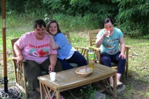 Na tle zieleni ławka ogrodowa, krzesło i stolik. Na ławce dwie śmiejące się młode kobiety. Jedna opiera się rękami o drugą. Obok nich siedzi dziewczyna, która pije coś z białego kubka. Na stoliku przed nimi stoi biały kubek jednorazowy, koszyk z chlebem i napój w butelce