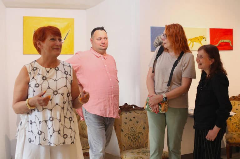 Wnętrze galerii. Grupa czterech osób prowadzi ożywioną rozmowę. Kobieta w prawej części kadru śmieje się. W tle kolorowe obrazy zawieszone na ścianie w jasnych barwach, w jednym rzędzie