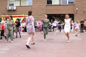 Na pierwszym planie trzy kobiety z personelu medycznego skaczące na skakankach. Dwie kobiety roześmiane, uchwycone w ruchu. W tle uczestnicy wydarzenia i ściana budynku szpitala