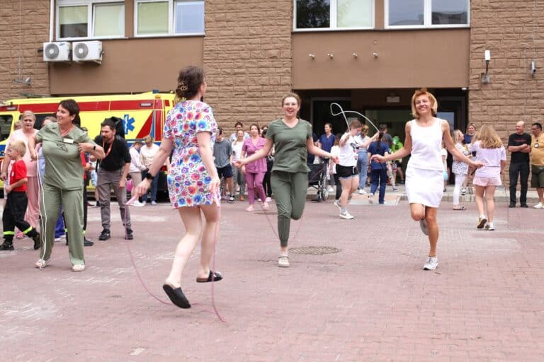 Na pierwszym planie trzy kobiety z personelu medycznego skaczące na skakankach. Dwie kobiety roześmiane, uchwycone w ruchu. W tle uczestnicy wydarzenia i ściana budynku szpitala