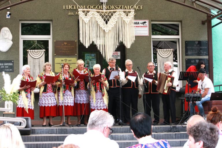 Na scenie grupa seniorów w lokalnych strojach: białe haftowane bluzki, czarne kamizelki i czerwone spódnice z koronkowymi białymi zapaskami. Mężczyźni w białych koszulach, czarnych kamizelkach i ciemnych spodniach