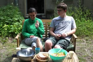 Na ławce siedzą od lewej: młoda kobieta w zielonej bluzce z długim rękawem, która macha do aparatu i chłopak w okularach, spodenkach i szarej koszulce. Przed nimi stolik, a na nim: metalowa miska, koszyk z chlebem, zielona miska z sałatką, kubki