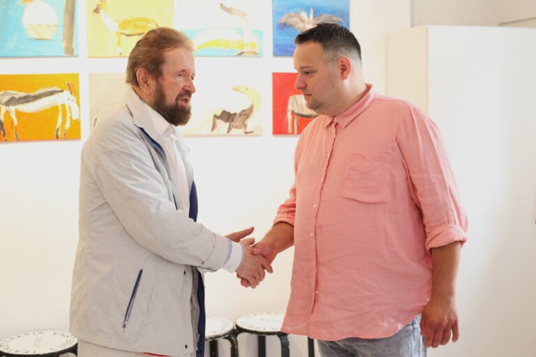 W centralnej części kadru Adam Saran przyjmuje gratulacje od gościa finisażu. Uchwycono moment uścisku dłoni obu mężczyzn. W tle, na ścianie w jasnych barwach wiszą w dwóch rzędach kolorowe obrazy