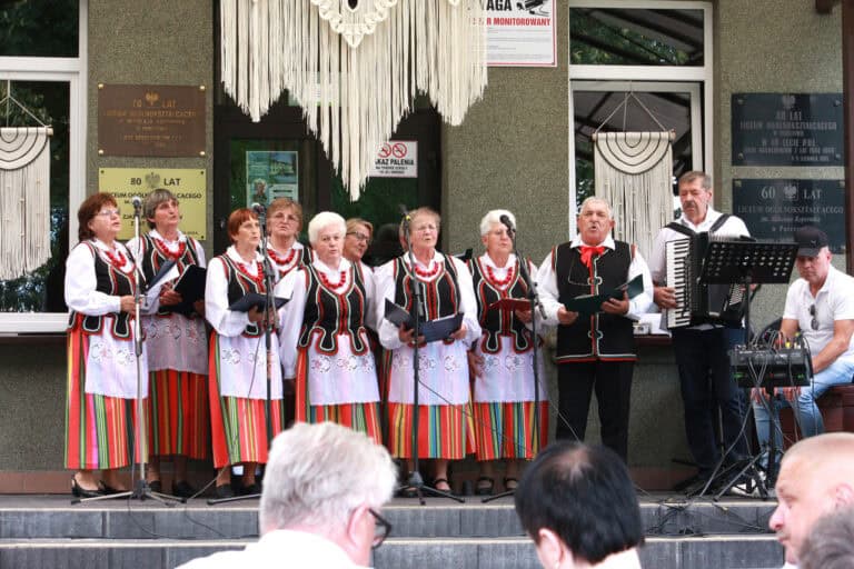 Grupa seniorów podczas występu. Kobiety ubrane w pasiaste spódnice z białymi zapaskami, białe bluzki i czarne kamizelki, wszystkie panie mają duże czerwone korale