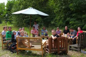 Grupa kilkunastu osób na ławkach i fotelach pod parasolem ogrodowym. Prawie wszyscy patrzą w kierunku aparatu fotograficznego. W tle zieleń