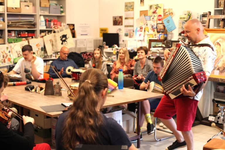 Wnętrz pracowni wypełnione uczestnikami warsztatów. Ostrość na lewą część zdjęcia. Na niej grający na akordeonie Marek Fedor
