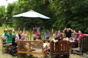 Grupa kilkunastu osób na ławkach i fotelach pod parasolem ogrodowym. Część osób patrzy z uśmiechem w kierunku obiektywu, unosząc w górę ręce. W tle zieleń drzew
