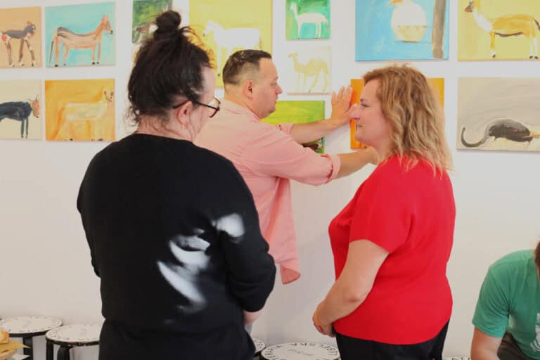 Na pierwszym planie dwie kobiety pogrążone w rozmowie. W tle Adam Saran poprawiający na ścianie obraz swojego autorstwa