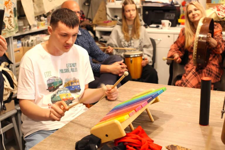 Zbliżenie na chłopca grającego na kolorowych cymbałach. W tle uczestnicy warsztatów i goście z Urzędu Miasta