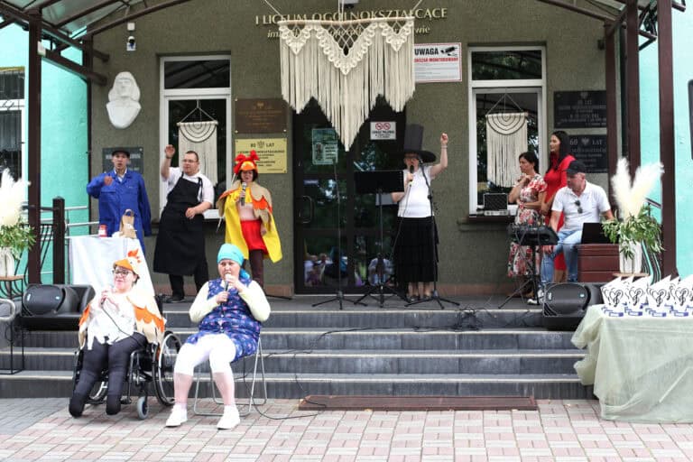 Na scenie artyści kabaretowi. Na pierwszym planie dwie siedzące kobiety (jedna z nich na wózku). Na drugim planie artyści w różnych strojach scenicznych wykonują gesty do publiczności. Jest wśród nich kobieta w wysokim cylindrze na głowie i druga w przebraniu kaczki, w kolorowym stroju imitującym skrzydła