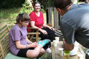 Dwie nastolatki w fotelach ogrodowych. Jedna z nich ma na sobie czerwoną bluzkę w białe poziome paski i uśmiecha się. Przed nimi stolik z napojami i białymi jednorazowymi kubkami