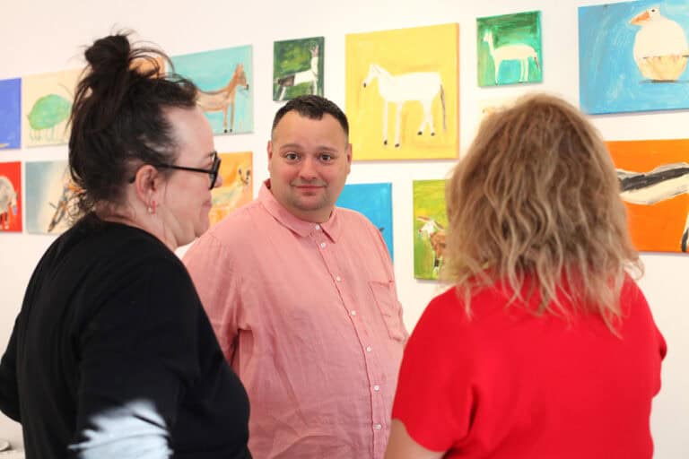 Na pierwszym planie dwie kobiety stojące tyłem do obiektywu. Pomiędzy nimi Adam Saran, uśmiechnięty, patrzy w obiektyw. W tle kolorowe obrazy artysty