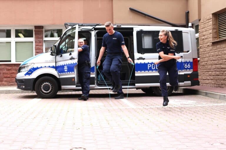 Grupa policjantów na tle policyjnego wozu. Jedna kobieta i dwóch mężczyzn. Na pierwszym planie kobieta i mężczyzna skaczą na skakankach
