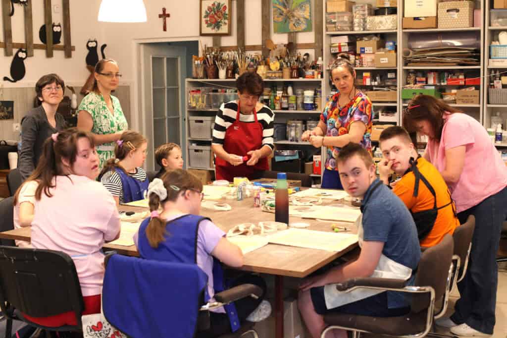 Przy dużym, kwadratowym stole siedzą dzieci i młodzież - uczestnicy projektu. Przy dzieciach pięć pań wspomagających. Część osób jest pogrążona w pracy, kilka osób patrzy w obiektyw. W tle regał w akcesoriami do malowania