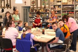 Przy dużym, kwadratowym stole siedzą dzieci i młodzież - uczestnicy projektu. Przy dzieciach pięć pań wspomagających. Część osób jest pogrążona w pracy, kilka osób patrzy w obiektyw. W tle regał w akcesoriami do malowania