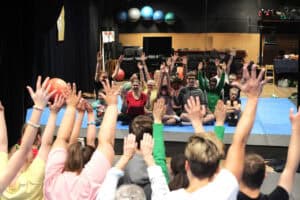 Odbicie w lustrze grupy osób. Wszyscy patrzą przed siebie i z uśmiechem unoszą w górę obie ręce. Siedzą na niebieskiej macie. W tle krzesła ustawione jedno na drugim, a nad nimi półka z dużymi piłkami w barwach: błękitu, szałwii i perłowej