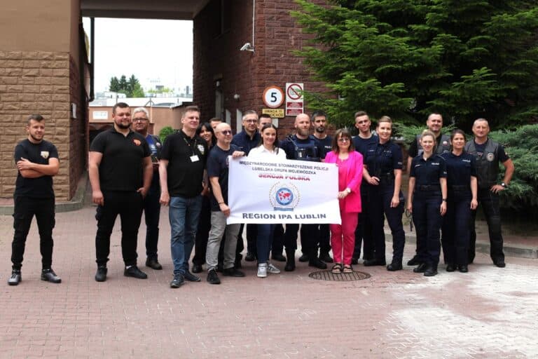 Grupowa fotografia, na której znajduje się kilkanaście osób. Klika z nich na pierwszym planie trzyma transparent z napisem: Międzynarodowe Stowarzyszenie Policji. Lubelska Grupa Wojewódzka. Sekcja Polska. Region IPA Lublin