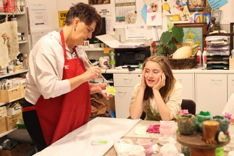 20. W centralnej części zdjęcia dziewczynka, która z uśmiechem obserwuje jak opiekunka maluje figurkę ceramiczną. W tle komody z akcesoriami plastycznymi