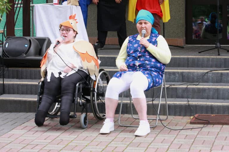 Dwie kobiety. Jedna z nich siedzi na wózku, druga na krześle. Obie mają w rękach mikrofony. Kobieta od lewej ma na sobie strój kury, druga ubrana w fartuch typu „podomka” i z niebieską chustką na głowie