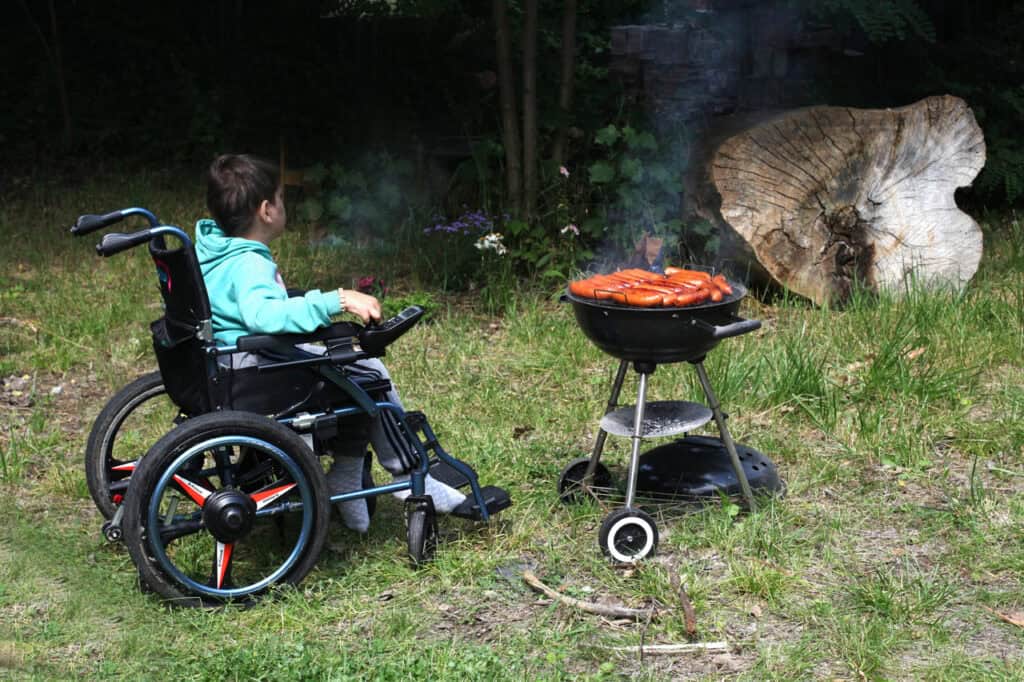 W centralnej części kadru grill z kiełbaskami. Z lewej chłopiec na wózku, wpatruje się w otaczającą roślinność. Po prawej pień uciętego drzewa